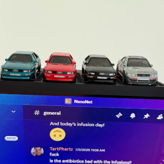 Four miniature die-cast model cars in teal, red, black, and silver, displayed on a black base above a computer monitor.