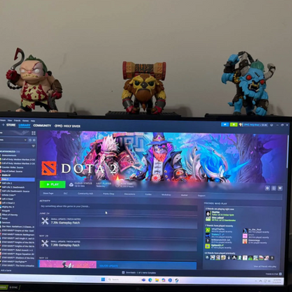 A computer monitor displaying the Dota 2 website, with three fantasy character figurines placed on top of the monitor against a plain wall.